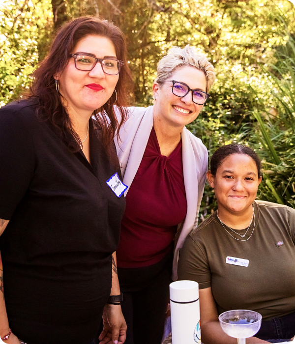 Three women are smiling together outdoors. The woman on the left has glasses and a name tag, the one in the middle has short blonde hair and glasses, and the one on the right is seated, wearing a green shirt with a name tag. Lush greenery is in the background.