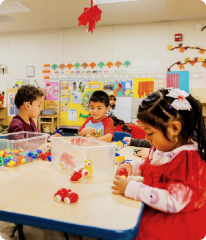 Four young children are seated around a table in a classroom, playing with colorful toys. The room has educational posters and student artwork on the walls. Curtains and decorations create a bright, engaging atmosphere.