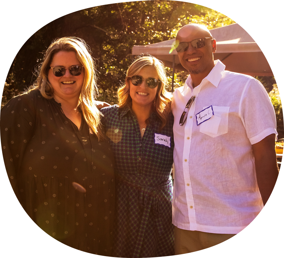 Three people, two women and one man, stand close together outdoors, smiling at the camera. They are all wearing sunglasses. The women have long hair and the man is bald. Each wears a name tag. Sunlight filters through trees behind them.