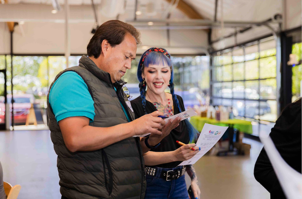 Two people standing inside a bright, indoor space. The person on the left, wearing a green vest over a blue shirt, is looking at a paper with concentration. The person on the right, with blue hair in braids, is smiling and holding a paper and pencil.