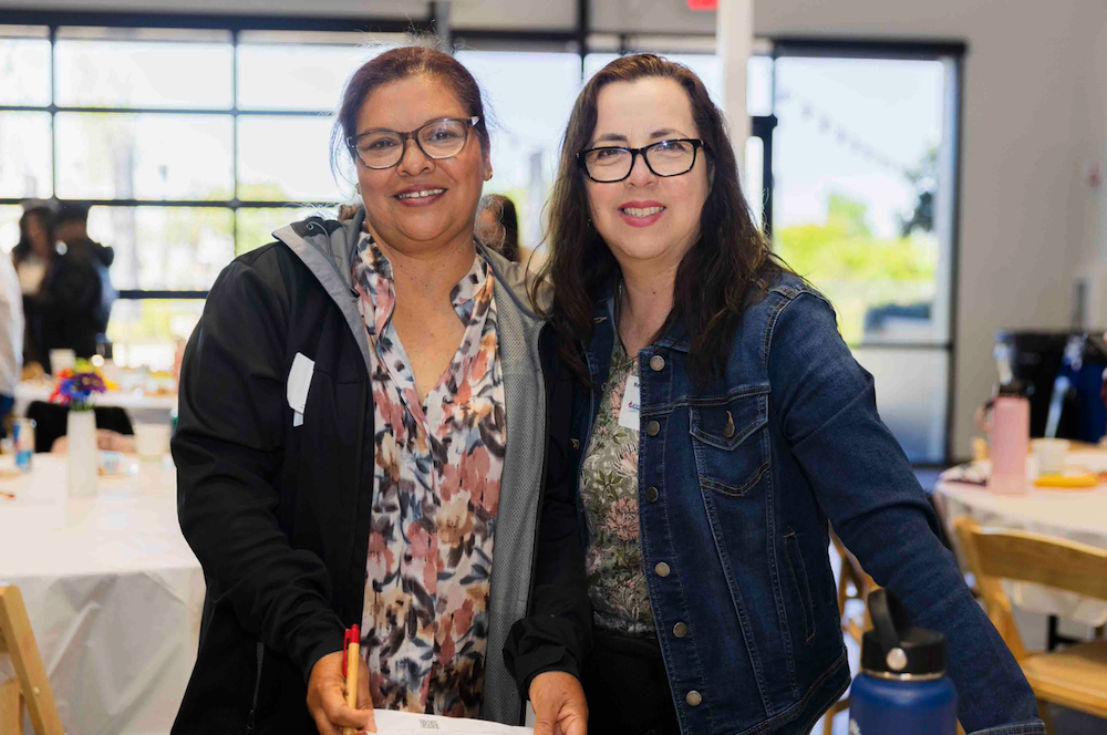 Two women, both with glasses, are standing side by side and smiling at the camera. One is wearing a black jacket and holding a piece of paper; the other is in a denim jacket. They appear to be in a well-lit room with tables and chairs in the background.