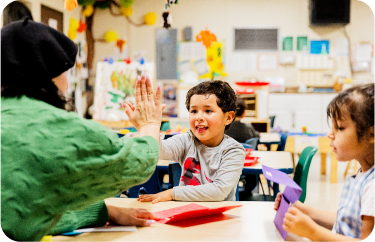 A child enthusiastically gives a high-five to an adult wearing a green sweater while sitting at a classroom table. Another child sits nearby, looking on with a smile. The classroom is colorful and filled with various crafts and educational materials.