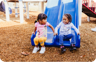 Two young children sit together at the bottom of a blue slide in an outdoor playground. One child is wearing a pink shirt and yellow pants, while the other is dressed in a white shirt and blue pants. Both are smiling and appear to be enjoying their time.