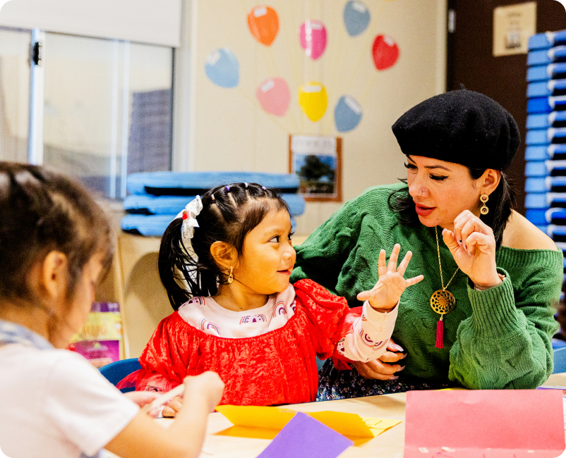 A woman in a green sweater and black beret engages with a young girl in a red dress at a classroom table. The girl looks up, showing her fingers, while another child on the left focuses on colorful paper. The background features wall decorations and classroom supplies.