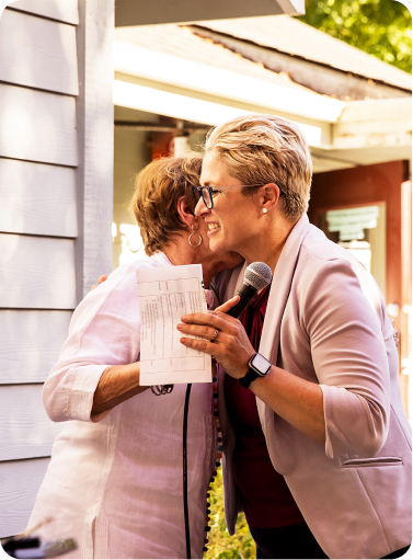 A woman with short blonde hair, wearing a light pink blazer, glasses, and a watch, is holding a microphone and a sheet of paper while hugging an older woman with short brown hair in a white shirt. They are outside a building, smiling warmly.