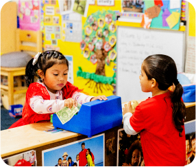 Two children are playing at a pretend store, with one child giving money to the other. The children are in a colorful classroom with a variety of educational posters, photos, and crafts displayed on the yellow walls.