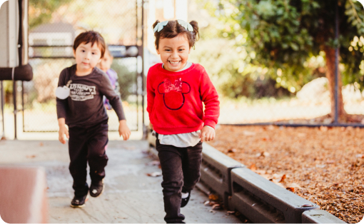 Two children are running outside. The child in the foreground, wearing a red sweater with mouse ears, is smiling widely. The child in the background, dressed in dark clothing, is also running and appears to be laughing. They are near a playground area with trees.