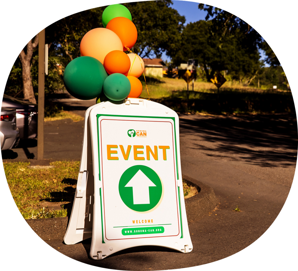 A white event sign with a green arrow, orange and green balloons attached, stands in a parking lot. The sign includes the logo "CAN" and directs attendees to the event. Trees and parked cars are visible in the background.