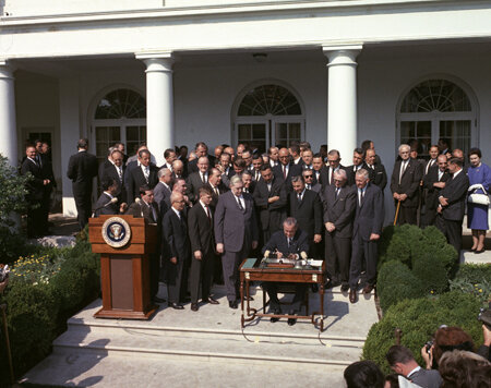 A historical outdoor gathering with a large group of men in suits. A person is seated at a small desk in front of the crowd, signing a document. A presidential podium is nearby, and the event takes place in front of a building with columns.