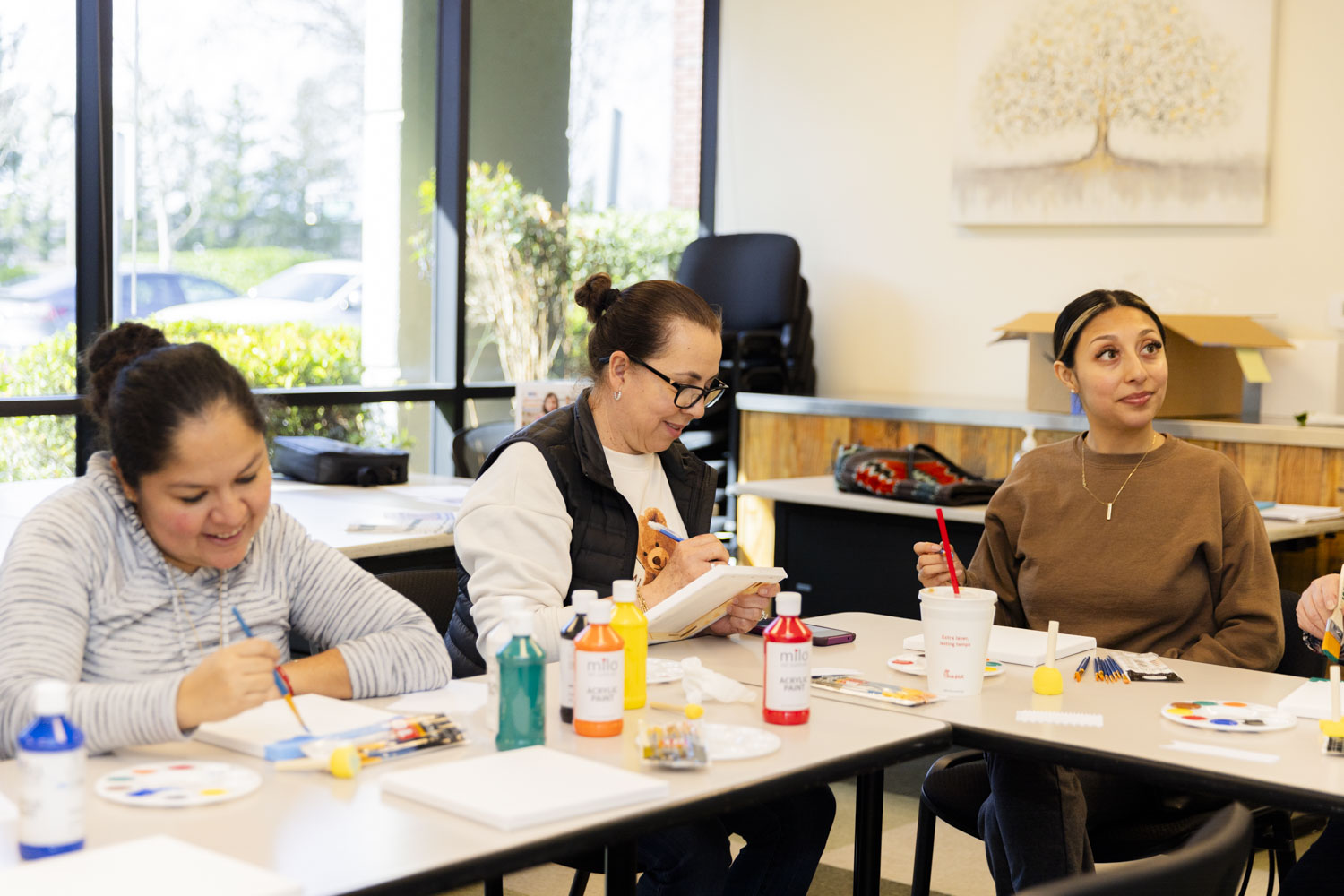 Four women sit at a table engaged in an art activity. They have paints, brushes, and canvas in front of them. The room is bright with large windows, and there's artwork on the wall. They appear focused and involved in the creative process.