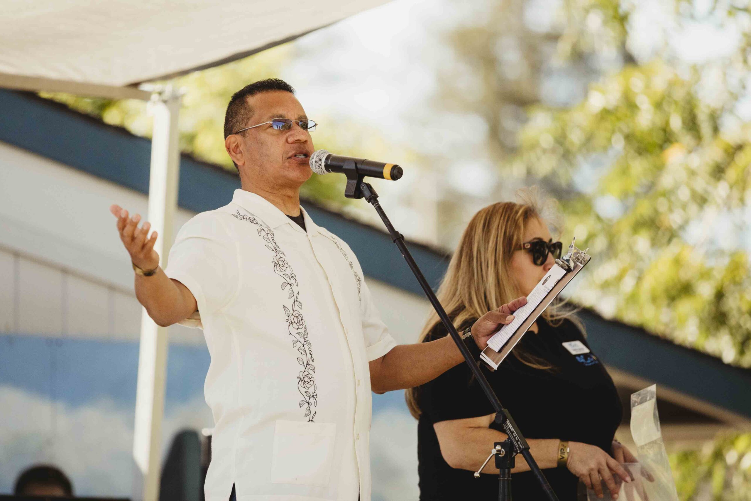 A man speaks into a microphone on an outdoor stage, wearing a white shirt with embroidered details. A woman beside him holds a clipboard and wears sunglasses. The background shows trees and a building under a partly cloudy sky.