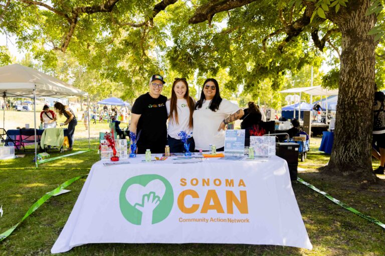 Three people stand behind a table with "Sonoma CAN Community Action Network" on the front. The table is outdoors on grass under a tree, displaying pamphlets and items. A small gathering is visible in the background with tents and people.