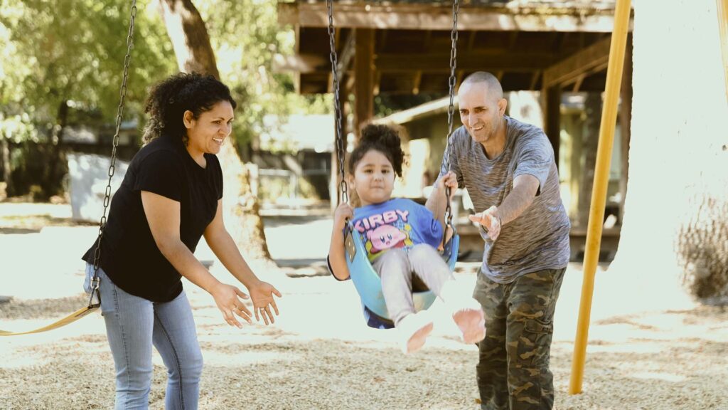 Two adults, a woman and a man, push a child on a swing in a playground. The child is smiling and wearing a shirt with a cartoon character. Trees and playground equipment are visible in the background.