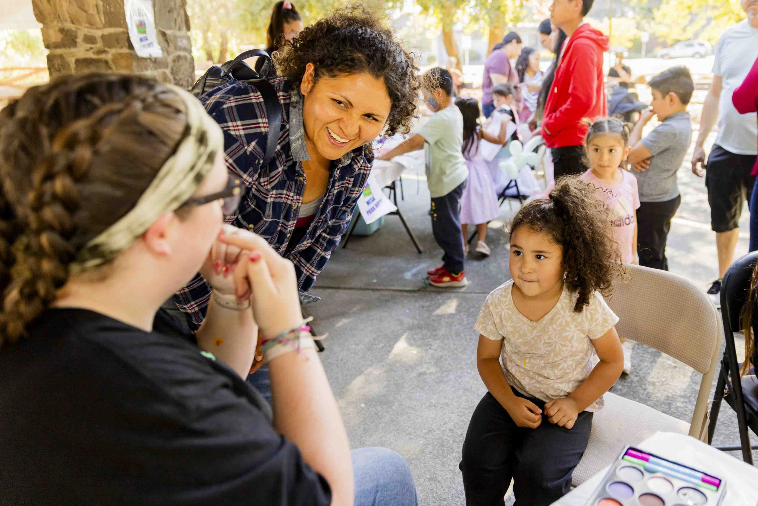 A woman smiling and talking to a seated woman, while a young girl sits nearby at an outdoor event. In the background, there are other people gathered, tables with items, and trees providing shade.