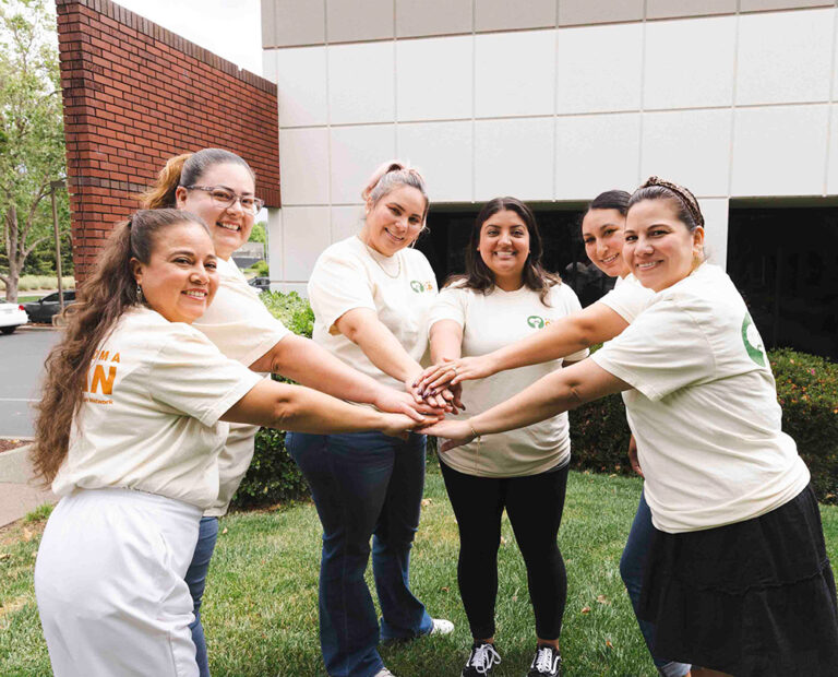 A group of six women wearing matching shirts stand in a circle outdoors, each with one hand in the center. They are smiling, with a building and trees in the background.