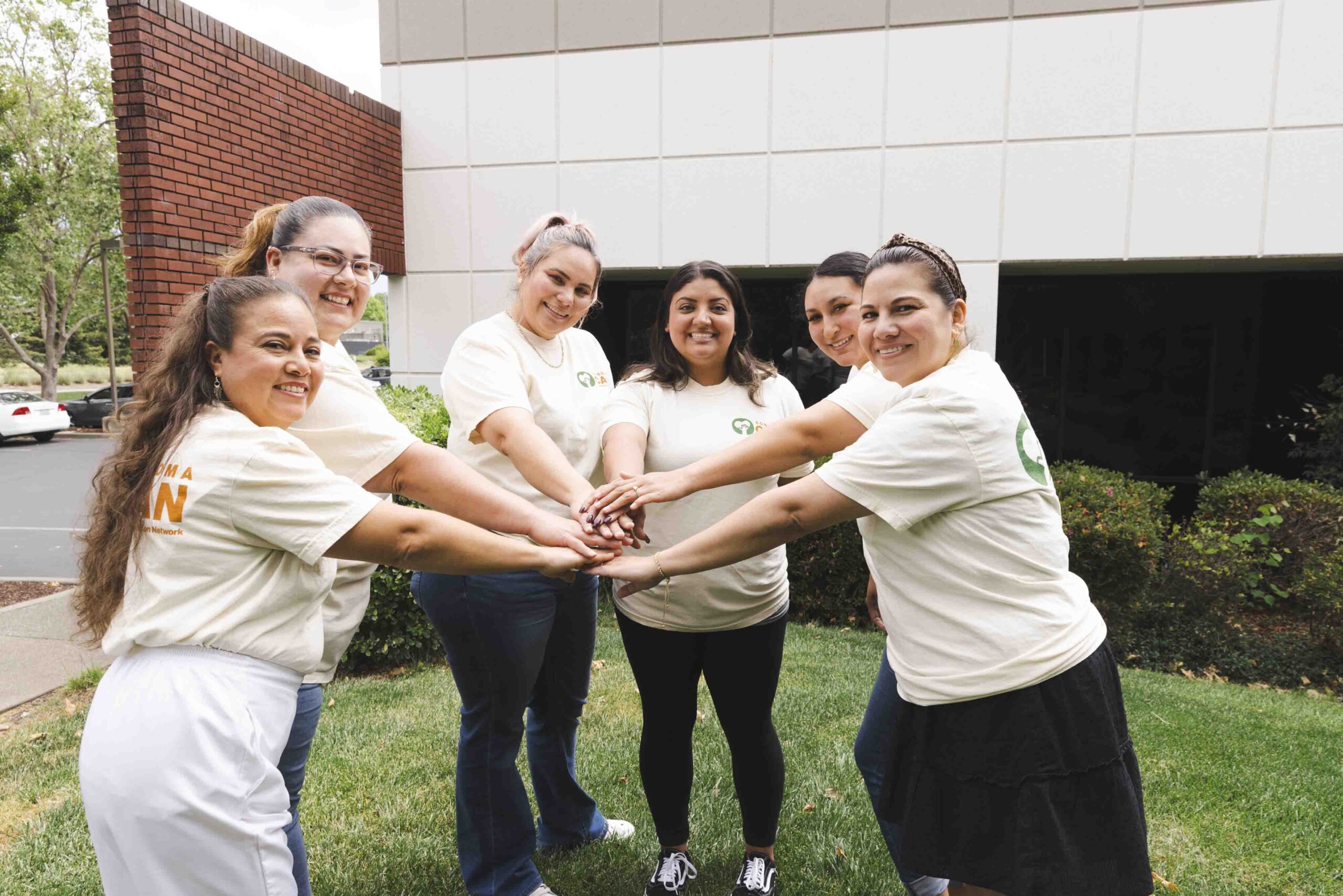 A group of six women, smiling and standing in a circle, stack their hands together in the center. They are outside on a grassy area, wearing matching light-colored shirts, in front of a building.