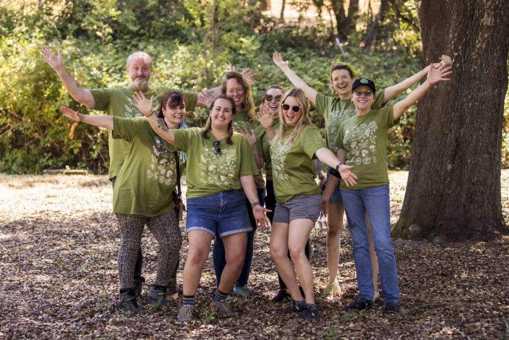 A group of eight people smiling and posing with arms outstretched in a wooded area. They are all wearing green shirts with nature-themed designs, suggesting a community or environmental event. Sunlight filters through the trees.