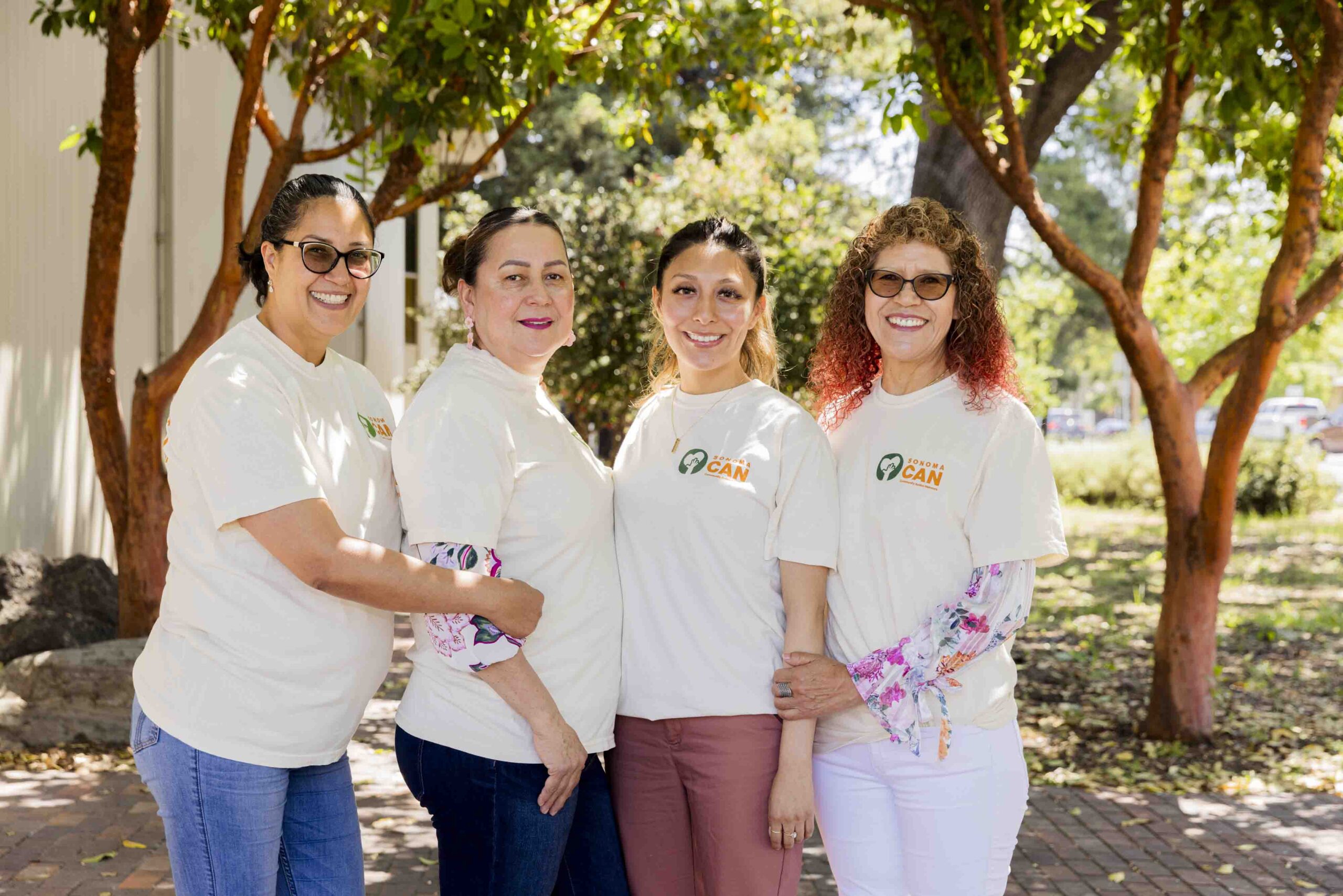 Four women stand together outdoors, smiling under trees. They wear matching T-shirts with a logo, and the setting is sunny and leafy, suggesting a park or garden environment.