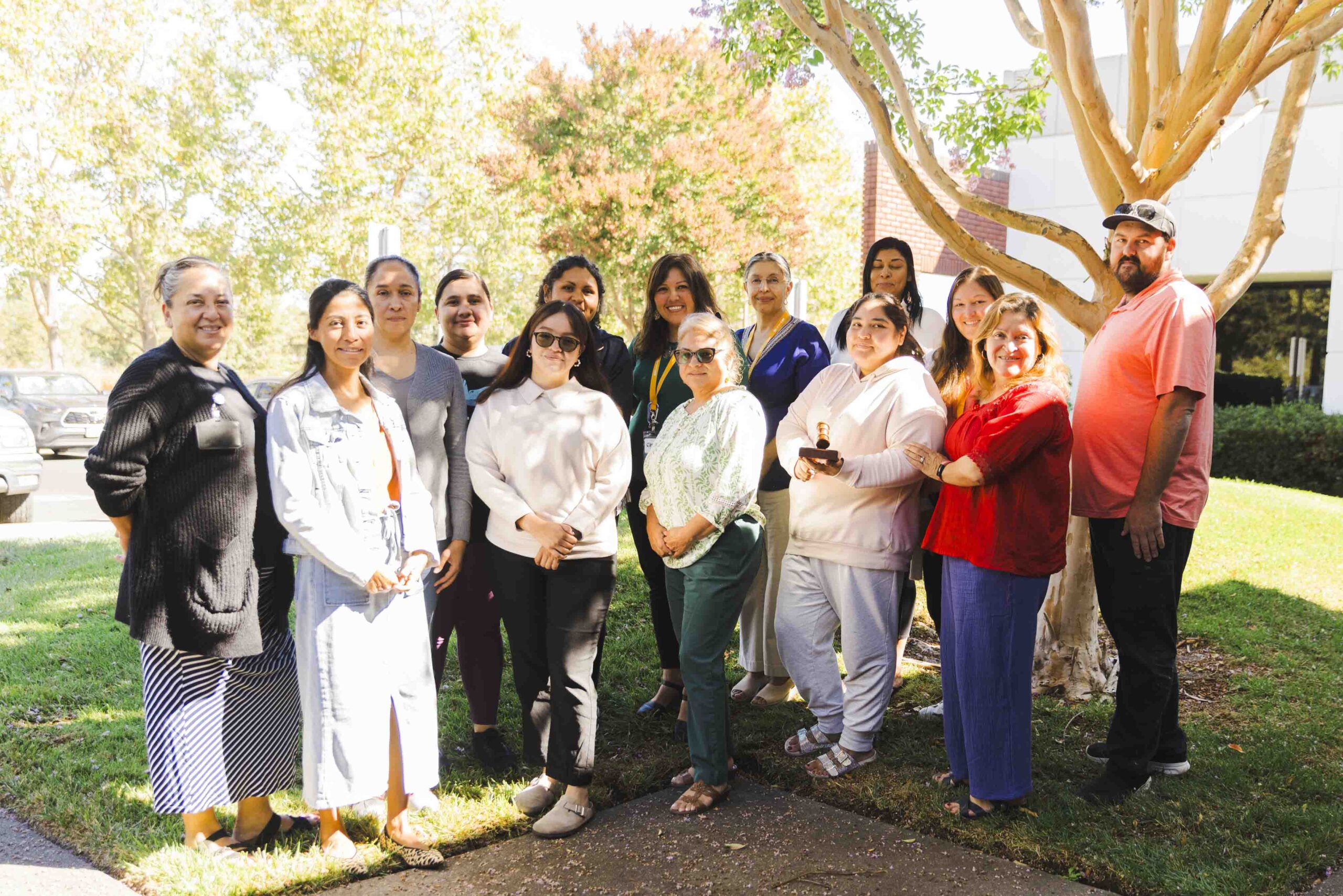 A diverse group of people posing outdoors in a park-like setting. They are standing on grass with trees and a building in the background, dressed in casual attire, smiling at the camera on a sunny day.