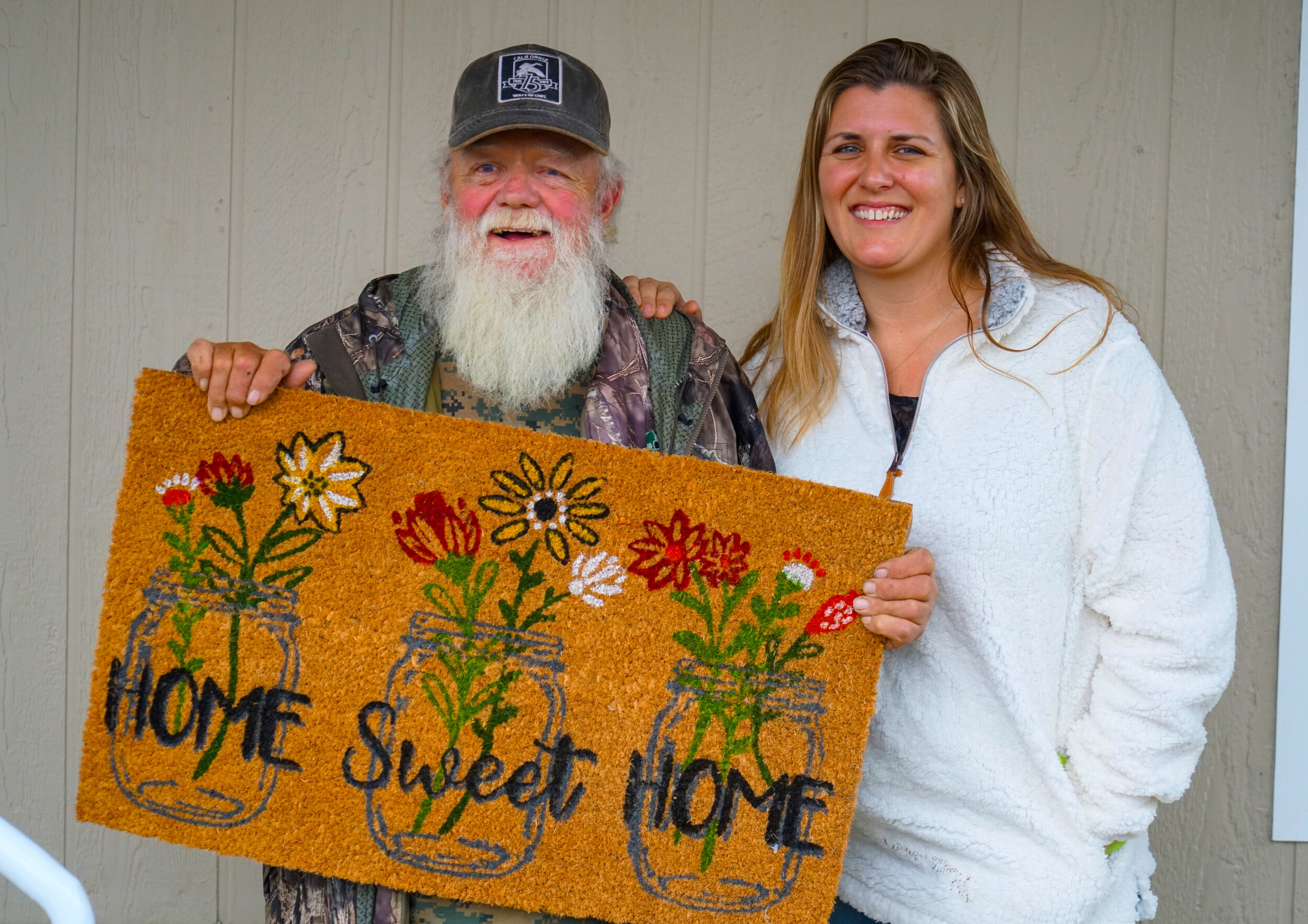 A smiling older man with a beard and a hat holds a "Home Sweet Home" doormat decorated with flowers. Beside him, a smiling woman in a white fleece jacket stands. They are both in front of a light-colored wall.