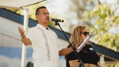 A man speaks into a microphone on an outdoor stage, wearing a white shirt with embroidered details. A woman beside him holds a clipboard and wears sunglasses. The background shows trees and a building under a partly cloudy sky.