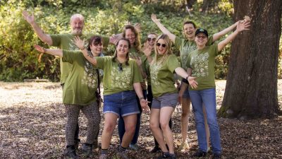 A group of eight people smiling and posing with arms outstretched in a wooded area. They are all wearing green shirts with nature-themed designs, suggesting a community or environmental event. Sunlight filters through the trees.