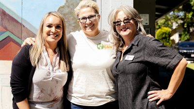 Three women are smiling and standing together outdoors. One wears a white T-shirt with a logo, the others wear casual tops. They are in front of a mural, and sunlight is casting shadows around them.